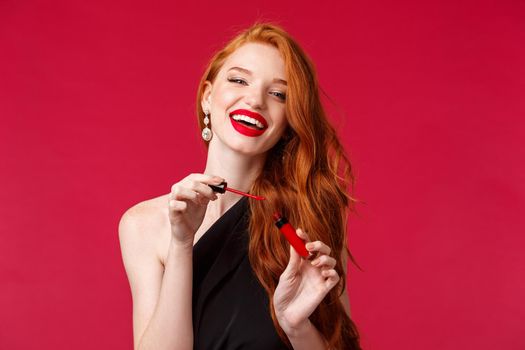 Makeup, beauty and women concept. Joyful good-looking feminine young woman with ginger hair, long curly hairstyle laughing, applying red lip gloss, do makeup before party, night-out.