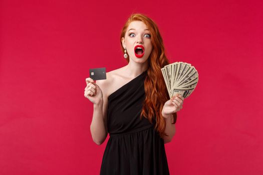 Luxury, beauty and money concept. Portrait of lucky and amazed girl express disbelief as receiving prize, thousands of dollars, hold fan of cash and credit card, look surprised, red background.