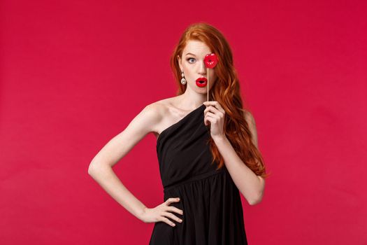 Celebration, holidays and women concept. Portrait of sassy and feminine young redhead woman in stylish black dress, holding lips card over eye and folding own in mwah kiss sign, red background.