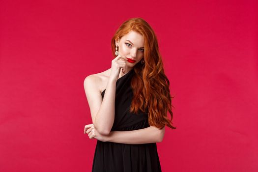 Portrait of sassy and seductive young elegant woman with red ginger hair, wearing black dress, evening makeup and red lipstick, look from under forehead daring and flirty, red background.