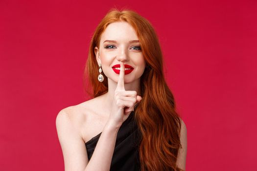 Close-up portrait of dreamy and pretty redhead woman with long curly hair hiding secret, prepared romantic surprise, shush with beaming smile, press index finger gently to lips, red background.