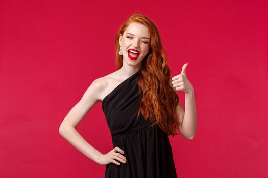 Fashion, luxury and beauty concept. Portrait of sassy gorgeous young redhead woman in black dress, wink at camera with thumb-up, recommend or approve something really awesome, likes party.