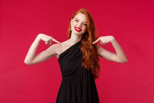 Fashion, luxury and beauty concept. Portrait of confident sassy redhead female in elegant black dress, wear makeup, pointing herself proudly, telling own achievement, red background.