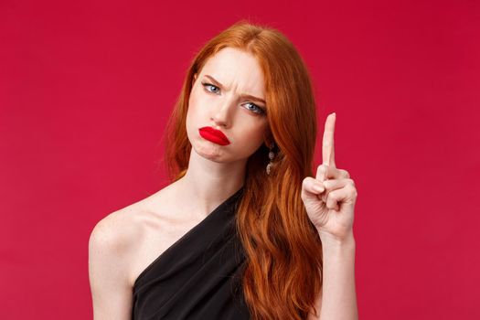 Close-up portrait of displeased moody redhead pretty girlfriend have one condition, frowning disappointed shaking finger as scolding someone, forbid or refuse, standing red background.