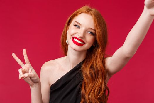 Close-up portrait of beautiful redhead european woman celebrating her birthday or enjoying party, taking selfie with peace sign and beaming satisfied smile, having fun, red background.