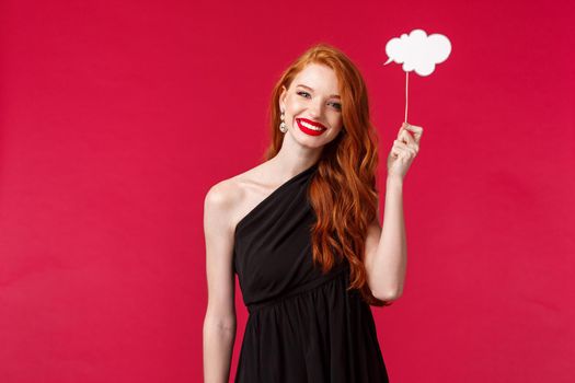 Portrait of charming elegant young redhead woman in black dress, holding comment cloud stick over head and smiling, searching for inspiration, have idea, stand red background.