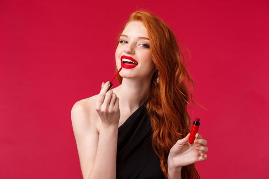 Makeup, beauty and women concept. Close-up portrait beautiful redhead feminine woman applying lipstick, red lip gloss and looking at camera as if in mirror, standing over red background.