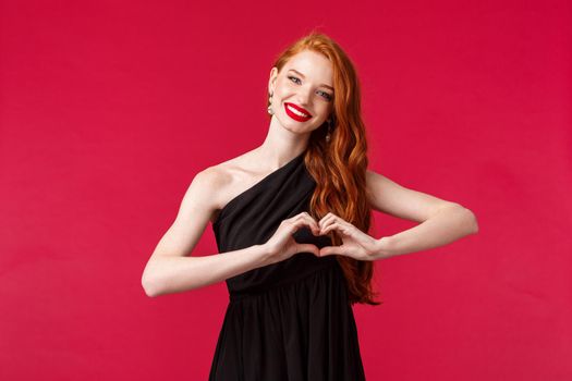 Portrait of charming elegant caucasian girl in red lipstick, black dress with red long hair, showing heart sign over chest and smiling heartwarming, express sympathy and appreciation, red background.