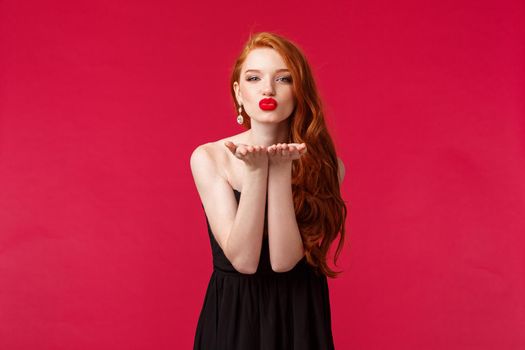 Portrait of coquettish feminine young beautiful redhead woman in black stylish dress, sending air kiss on palms near folded lips, looking at camera give sensual mwah, red background.