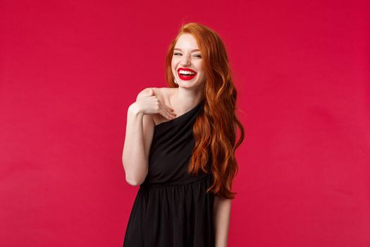 Portrait of elegant coquettish redhead woman in black dress and evening makeup, laughing silly at camera, gesturing as if talking to person during formal event, stand red background.