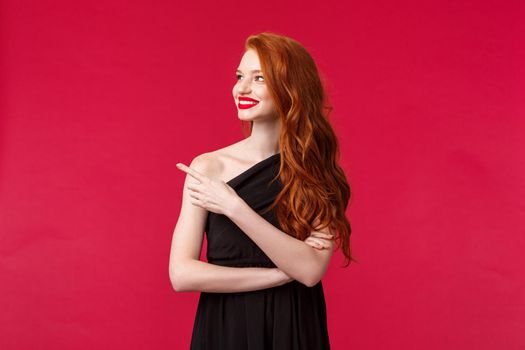 Celebration, events, fashion concept. Satisfied, charming redhead woman with red lipstick, wear black elegant dress, pointing and looking left with happy pleased expression, black background.