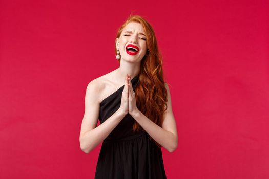 Portrait of gloomy desperate young redhead woman in black evening dress, whining and crying her heart out as pleading for forgiveness, close eyes sobbing, begging for help, red background.