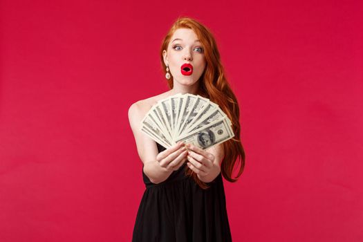 Luxury, beauty and money concept. Portrait of amazed and happy good-looking redhead woman showing winner his prize cash, giving dollars and praising with great bet, stand red background.