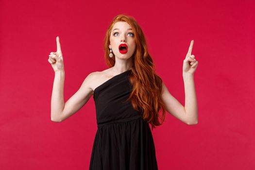Celebration, events, fashion concept. Portrait of amazed beautiful redhead woman in elegant evening dress, open mouth amused look and pointing fingers up at promo, red background. Copy space