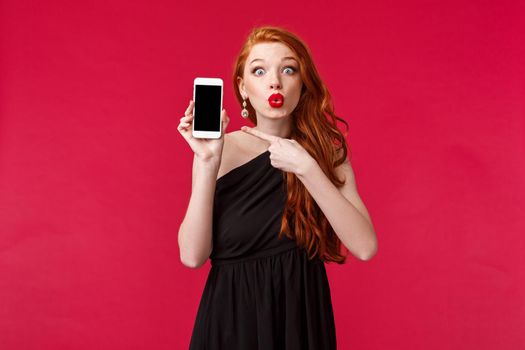Portrait of excited redhead girl with long ginger hair, wear black elegant dress, folding lips in amazement, talking about new app, or post from her crush social media, pointing mobile display.