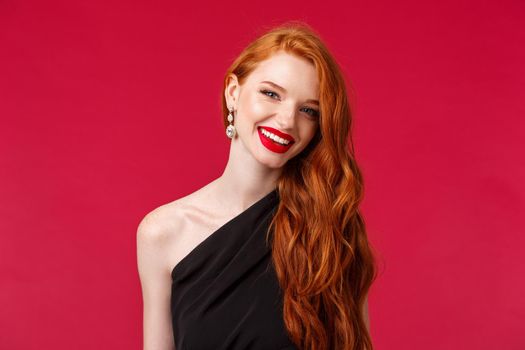 Close-up portrait of elegant gorgeous young redhead woman with red lipstick, evening makeup and earrings, wear black dress, have beaming smile on face, express joy and happiness.