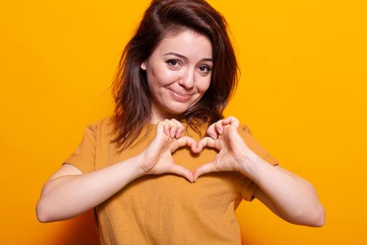 Positive adult making heart shape with fingers at camera. Portrait of romantic person doing love sign and symbol with hands, showing affection and emotion over orange background.