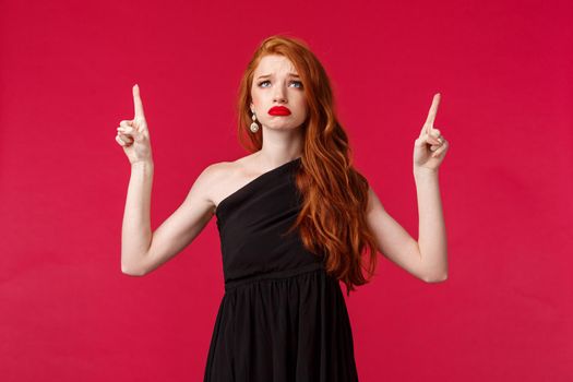 Celebration, events, fashion concept. Portrait of grieving gloomy, upset cute redhead woman in black dress, pointing fingers up, sighing and looking with regret and jealousy, dreaming of something.