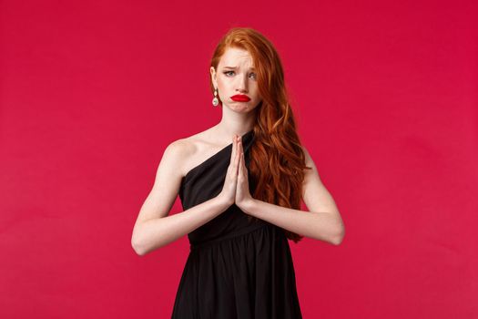 Portrait of gloomy and upset, sad redhead woman in black evening dress asking for help, need something asap, grimacing uneasy hold hands in pray, pleading or begging, stand red background.