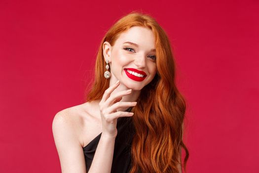 Close-up of happy gorgeous redhead woman with earrings, red lipstick and evening black dress, gently touching her jawline and beaming smile at camera cheerful, enjoying celebration, party concept.
