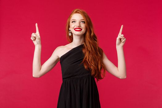 Celebration, events, fashion concept. Portrait of elegant and dreamy sensual redhead woman in black dress, pointing looking up with happy satisfied expression, found what she needs.