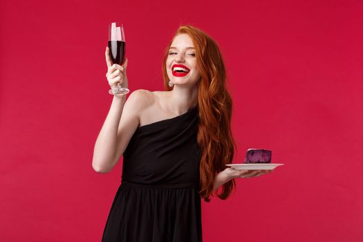Happy gorgeous and elegant feminine redhead woman raising glass of champagne saying toast or cheers, drinking celebrating birthday or having fun time at party in black dress, red background.