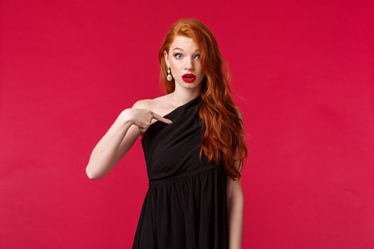 Fashion, luxury and beauty concept. Portrait of surprised and indecisive redhead caucasian woman in elegant black dress asking question, look confused, pointing herself with disbelief, red background.