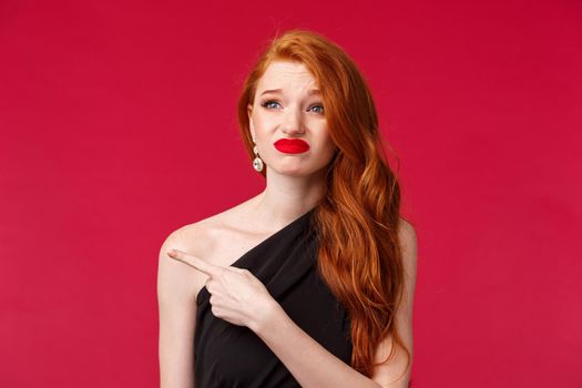 Close-up portrait of skeptical and indecisive cute grimacing redhead girl squinting and smirk as something painful, pointing looking finger left strange, cringy or weard thing, stand red background.