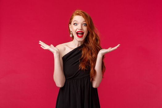 Fashion, luxury and beauty concept. Portrait of happy and surprised excited redhead woman react to unexpected pleasant guest showing-up her party, raise hands sideways and smiling, black dress.