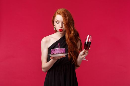 Silly good-looking redhead woman in slim black dress feel drunk after having few glasses champagne, hold glass and b-day cake, blowing out candle and making with during birthday party.