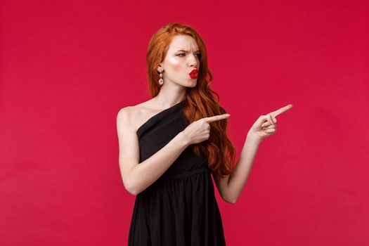 Celebration, events, fashion concept. Portrait of disappointed and complaining young sexy redhead woman in black dress, frowning grimacing frustrated, pointing looking right.