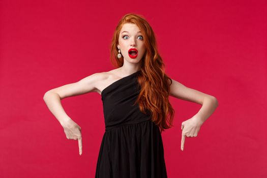Celebration, events, fashion concept. Surprised and amazed redhead young woman in black dress attend awesome event, talking about cool news, pointing fingers down and stare camera stunned.