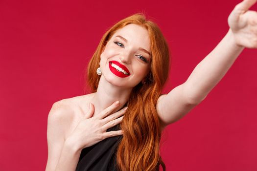 Close-up portrait of gorgeous smiling, happy redhead woman enjoying her birthday party, taking selfie for social media in stylish black dress and prom look with red lipstick, red background.