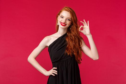 Fashion, luxury and beauty concept. Portrait of satisfied coquettish pretty redhead woman in elegant black dress, showing guarantee, okay sign with pleased nod, smiling happy, red background.