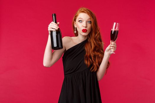 Celebration, holidays and women concept. Portrait of pumped and excited stylish pretty redhead woman suggesting have a drink, wear black dress, hold bottle of champagne and glass, partying.