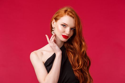 Close-up portrait of sensual seductive redhead woman with red lipstick, touching earring coquettish look camera, slightly smiling as flirting with person on party, wear black dress, red background.