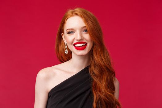 Close-up portrait of feminine gorgeous young redhead caucasian woman in red lipstick, earrings and evening slim black dress, smiling happy and pleased, having conversation with person on party.