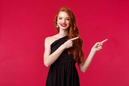 Celebration, events, fashion concept. Portrait of sensual happy gorgeous redhead woman in black slim elegant dress, pointing fingers right and smiling, wear makeup, black background.