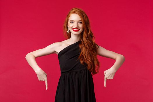 Celebration, events, fashion concept. Portrait of cheerful, smiling elegant young redhead woman in black dress, pointing fingers down and looking camera happy, advice click link, red background.