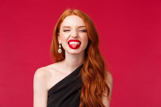 Close-up portrait of carefree, happy fabulous young redhead woman in black elegant dress, show tongue and silly smiling, squinting carefree enjoying party, celebrating holiday.
