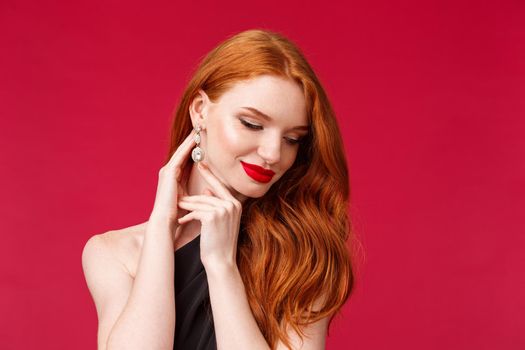 Close-up portrait of feminine tender and elegant young redhead woman with long curly ginger hair, earrings, tilt head look down with silly seductive smile, coquettish flirting, wear black dress.