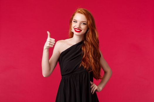 Fashion, luxury and beauty concept. Portrait of satisfied, assertive redhead elegant girl in black dress, show thumbs-up in approval or like gesture, smiling approvingly, red background.