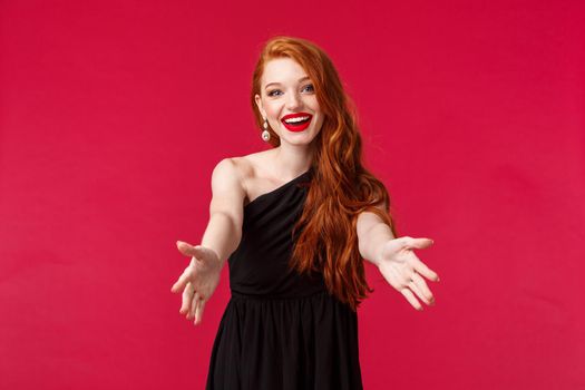 Fashion, luxury and beauty concept. Come here dear. Charming gorgeous redhead woman, ginger hair, wear black evening dress, reaching hands for hug, greeting guests to her party, welcome people.