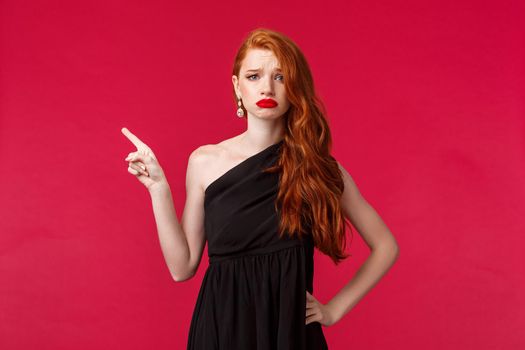 Beauty, fashion and women concept. Upset and displeased young gloomy redhead girl sighing and frowning unhappy pointing left look camera, express sadness wear black evening dress.