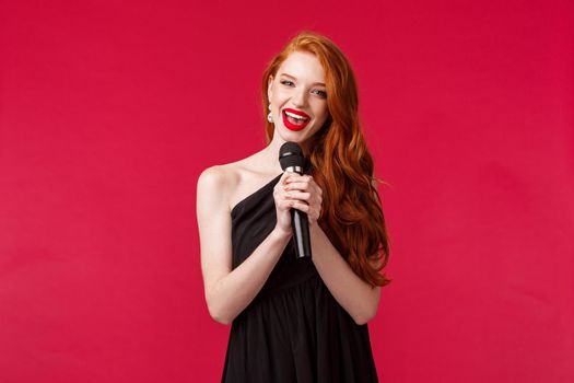Portrait of feminine gorgeous young woman in black dress, holding microphone singing karaoke or perform songs in front of audience on public event, standing red background.