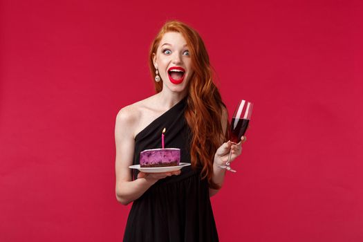 Portrait of excited and amused happy redhead b-day girl holding cake with lit candle and trying make wish blow it for dream come true, celebrating at party drinking wine, red background.