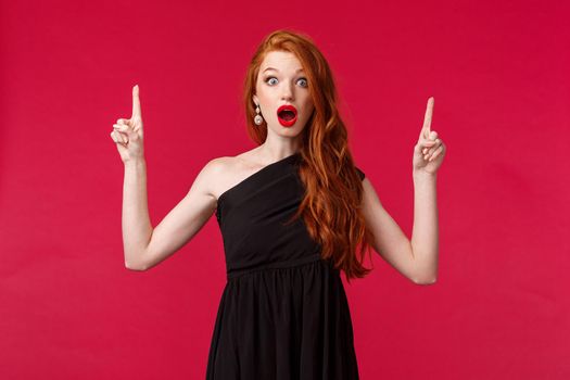 Celebration, events, fashion concept. Portrait of amazed and wondered, excited redhead woman in black dress, makeup, stare at camera impressed, pointing fingers up at advertisement.
