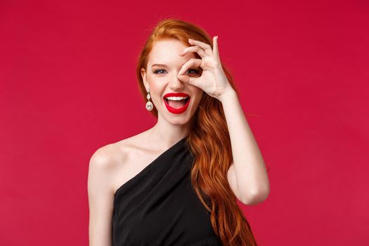 Close-up portrait of enthusiastic luxurious gorgeous redhead woman with long curly red hair, black dress and lipstick, look through okay sign and smiling joyfully, found something perfect.