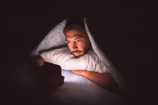 Young man lying in bed under a blanket and using smartphone at night. Social media addiction and communication. High quality photo