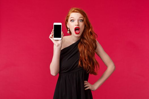 Portrait surprised and impressed redhead girl in black dress, drop jaw in amazement and shock, holding mobile phone, showing smartphone screen to friend and gossiping about persons profile.